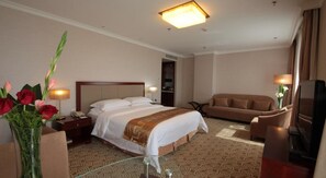 Quarto de hotel com cama, sofá, televisão e escrivaninha com luminária.