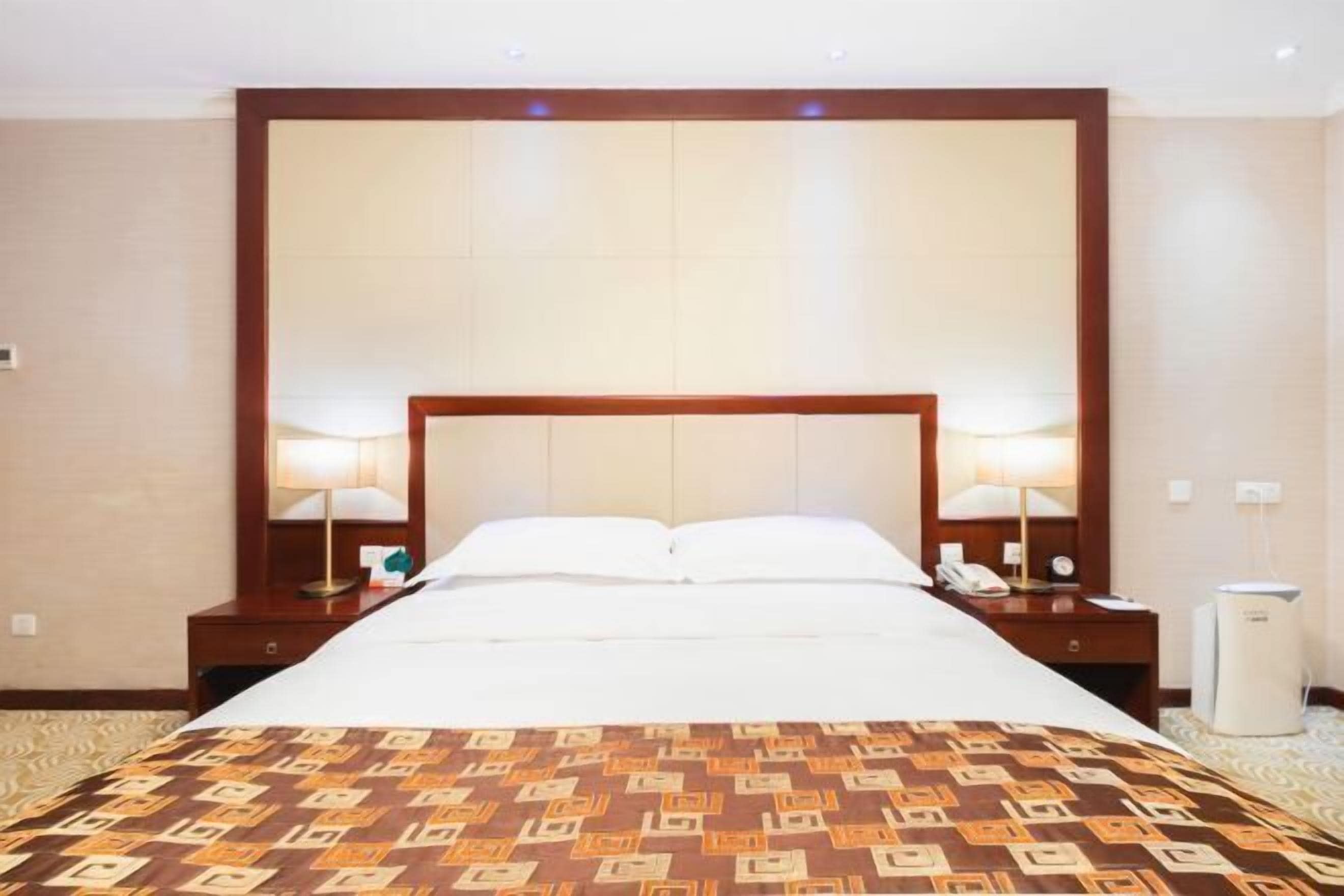 A hotel room with a large bed, two bedside tables, a nightstand, a lamp, a phone, and a heater.