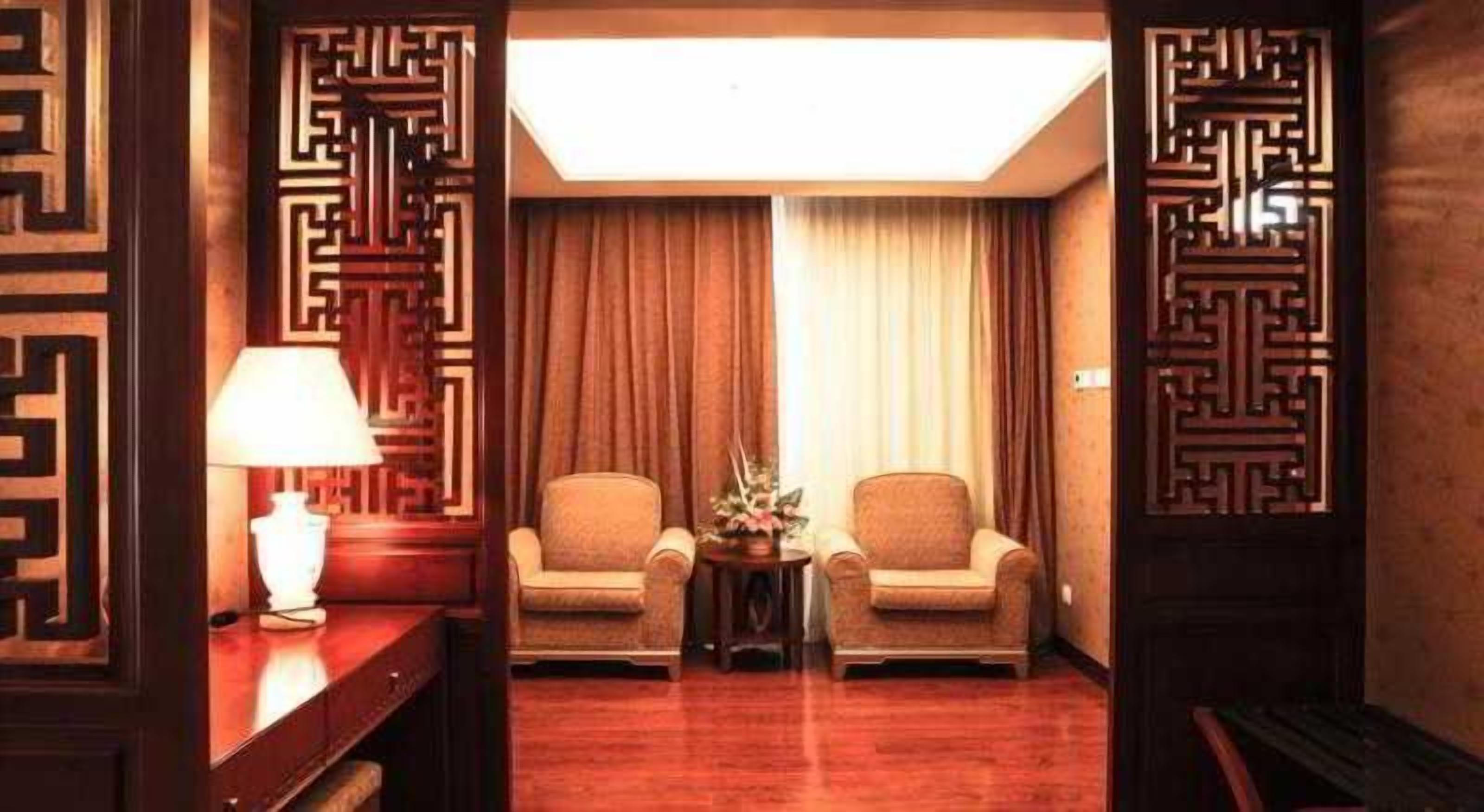A hotel room with a wooden floor, two armchairs, a small table with a lamp, and a decorative screen.