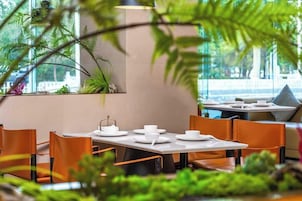 Comedor con mesas y sillas, rodeado de plantas de interior y vistas a través de amplios ventanales.