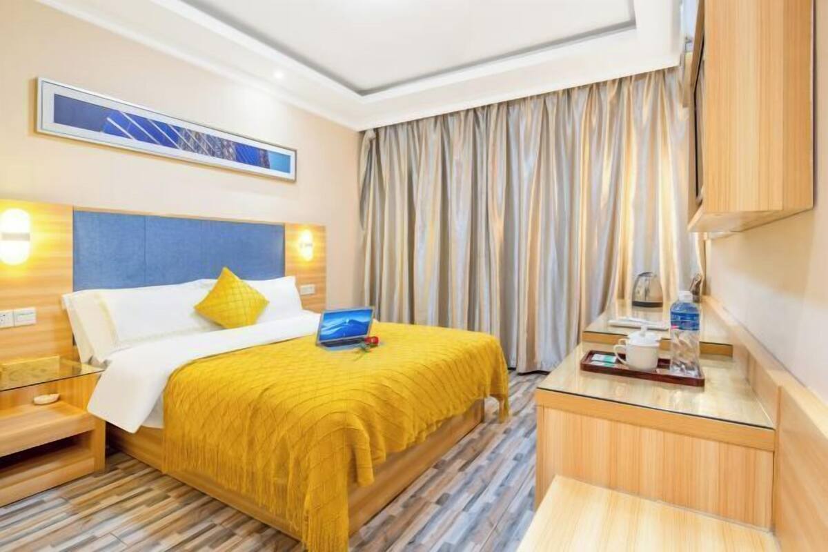 A hotel room with a bed, bedside tables, a desk with a chair, a window with curtains, and a wall-mounted TV.