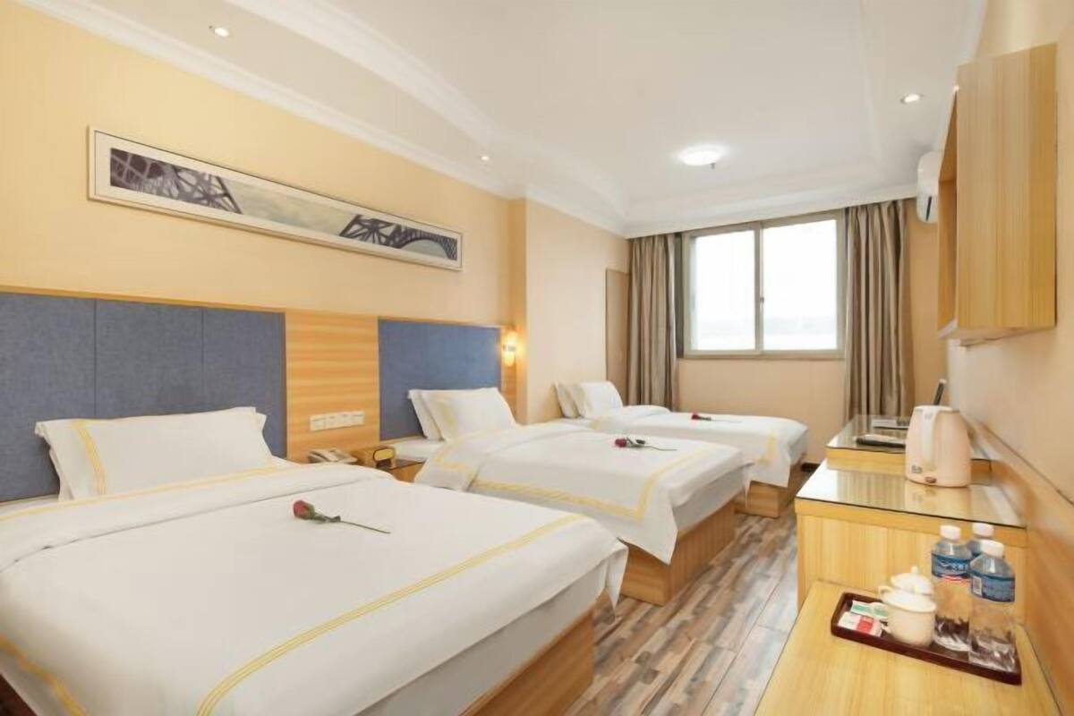 A hotel room with three beds, a wooden headboard, a window with curtains, a bedside table with a kettle, and a tray with toiletries.