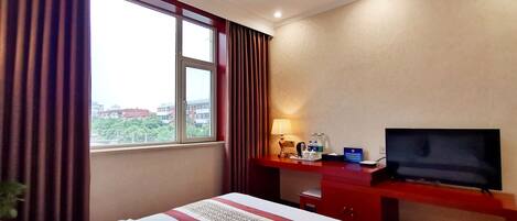 Quarto de hotel com cama, escrivaninha com televisão, luminária e janela com cortinas.