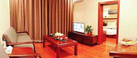 A hotel room with a wooden floor, a TV on a wooden stand, a bed with white bedding, a wooden cabinet, and a dining table with a floral centerpiece.