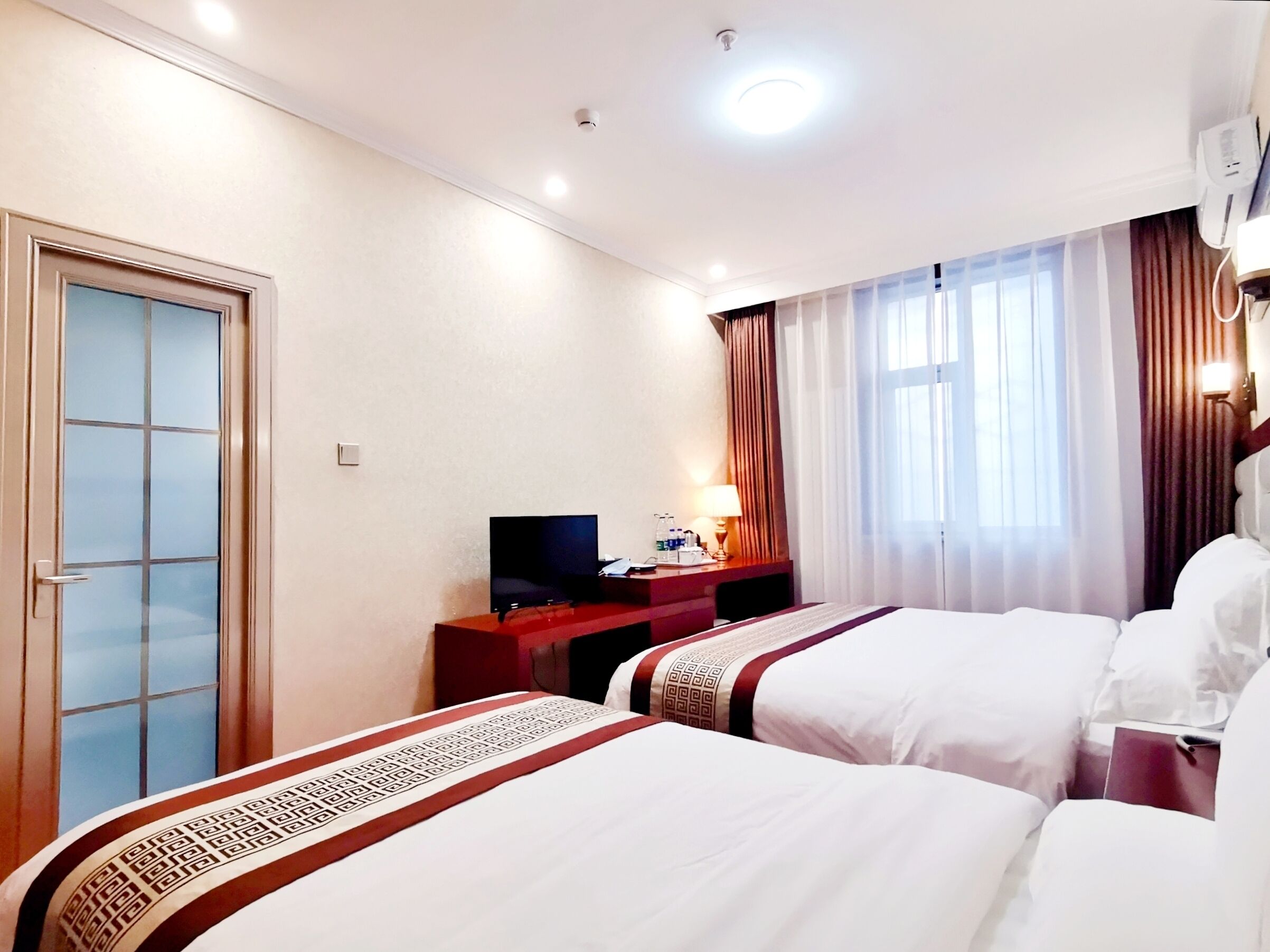 Quarto de hotel com duas camas, televisão, escrivaninha com candeeiro e janela com cortinas.