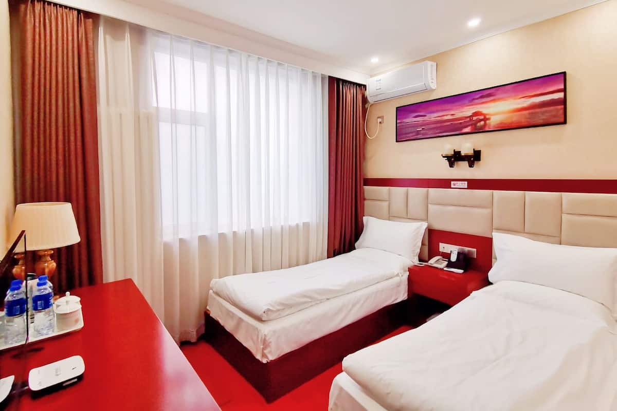 Une chambre d’hôtel avec deux lits, un bureau rouge, une lampe, une bouteille d’eau, une télécommande et un tableau accroché au mur.