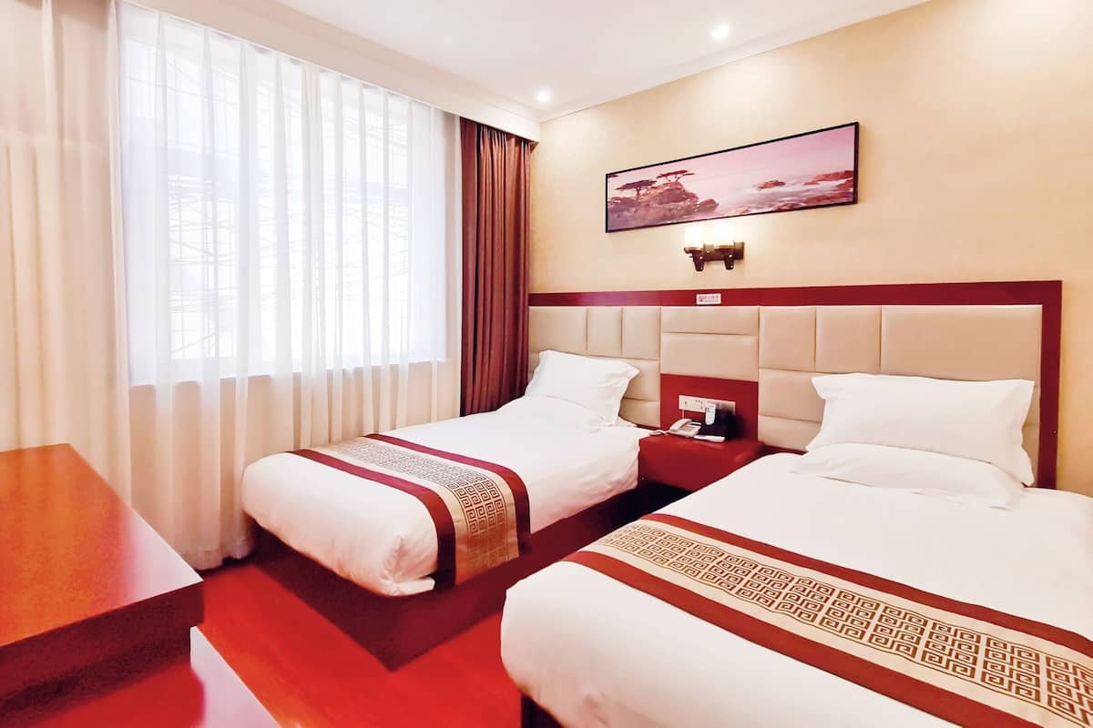 Une chambre d’hôtel avec deux lits, une grande fenêtre avec des rideaux légers, un tableau au mur et un mur d’accent rouge.