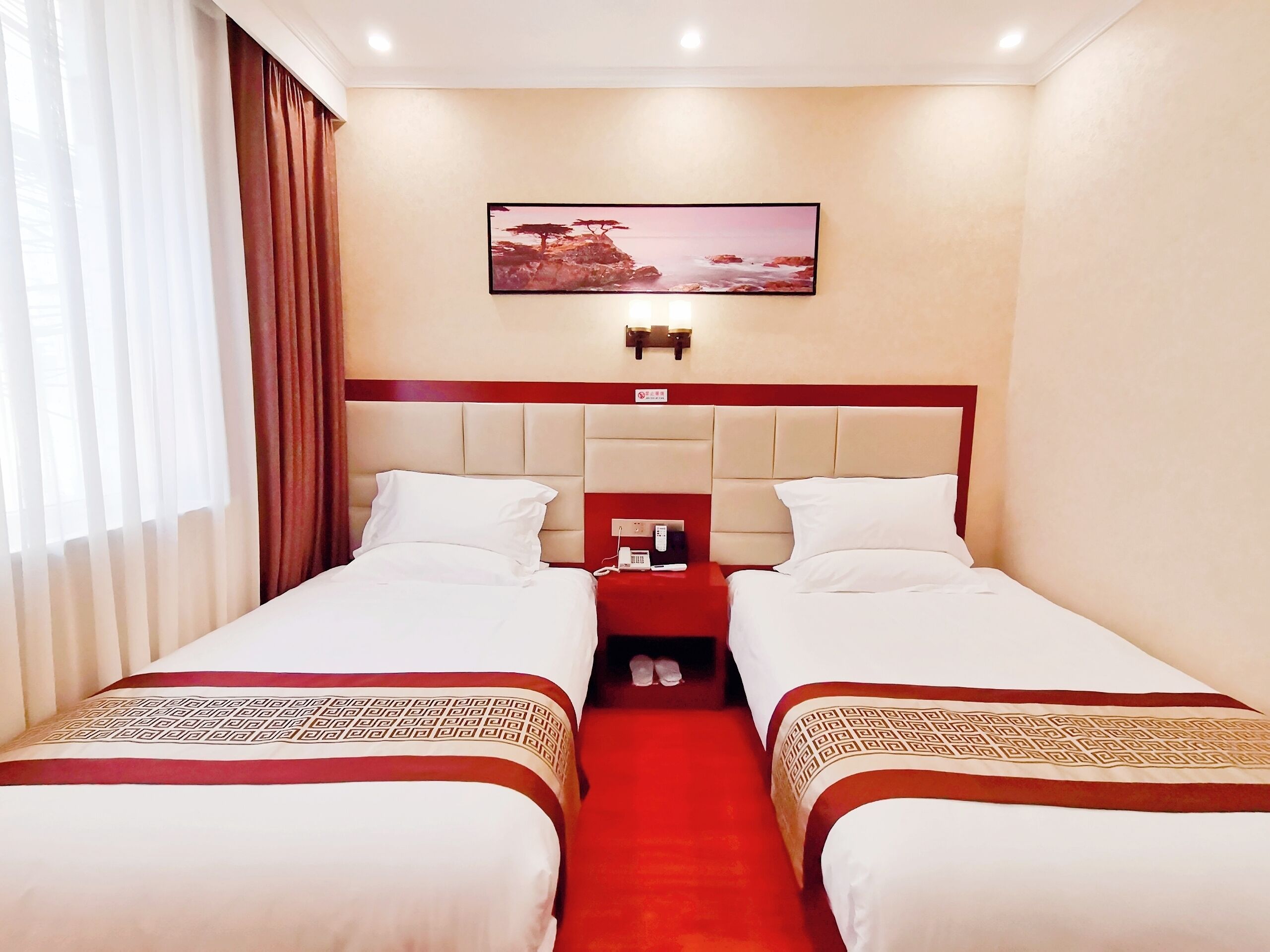 Habitación de hotel con dos camas, una alfombra roja, un cuadro en la pared y una mesita de noche con teléfono y cargador.
