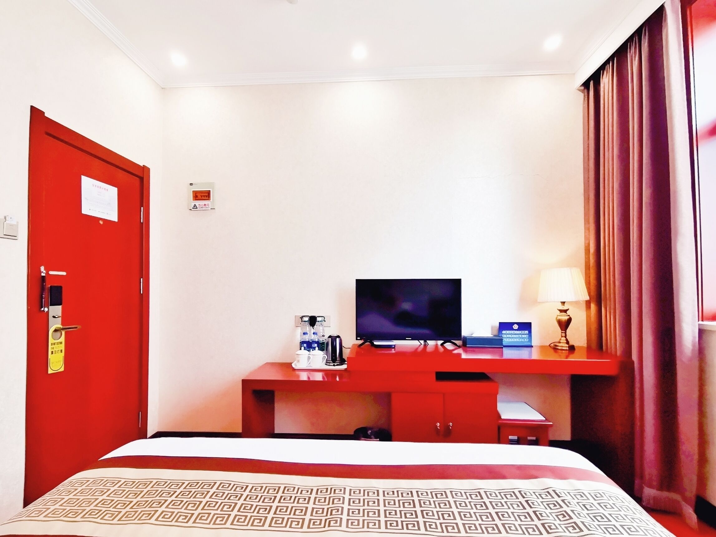 Ein Hotelzimmer mit einem Bett, einem Schreibtisch mit Fernseher, einer Lampe und einer roten Tür.