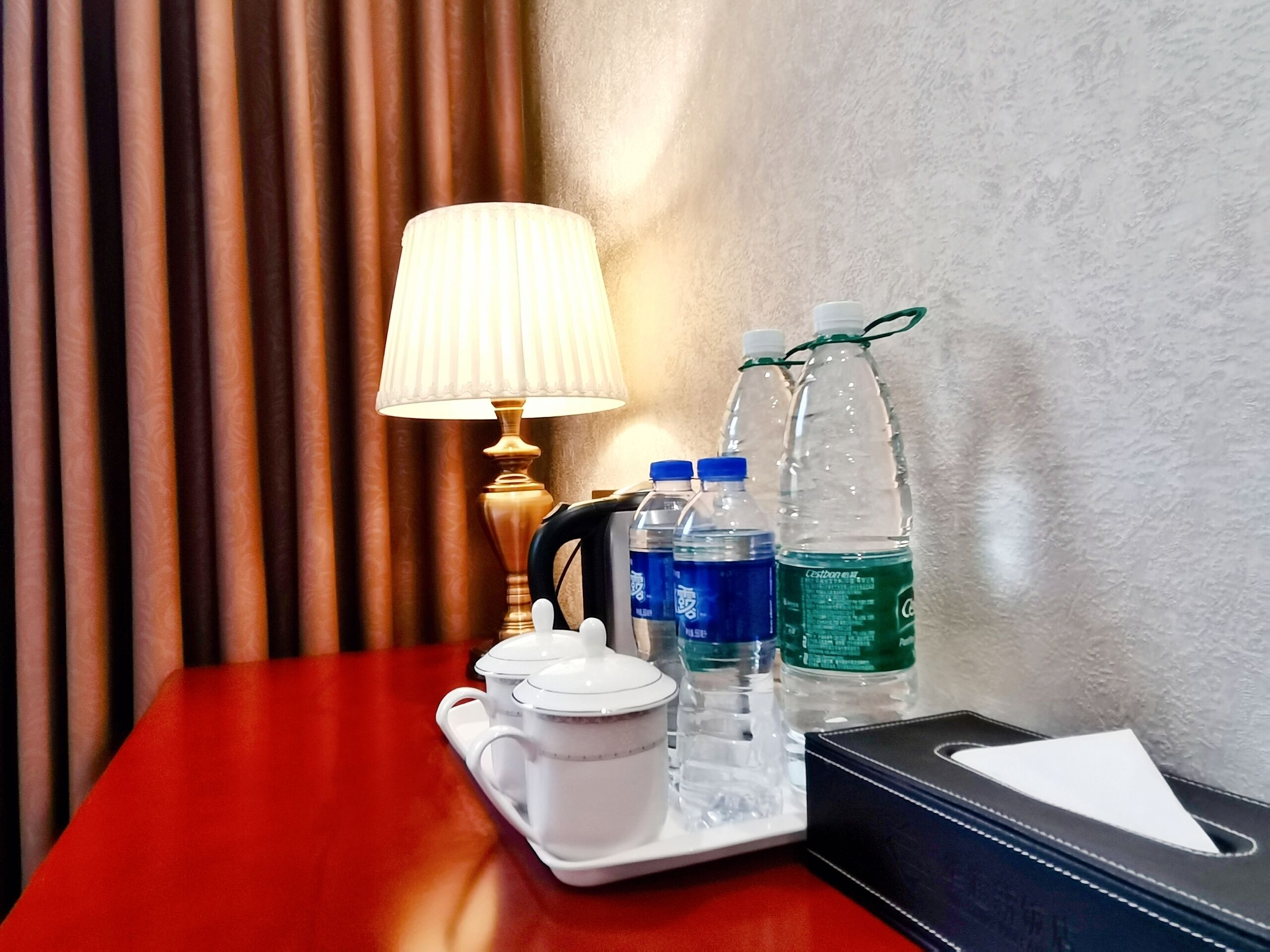 Habitación de hotel con una mesa de madera, una bandeja blanca con tetera y tazas, dos botellas de agua, una lámpara y una caja de pañuelos.
