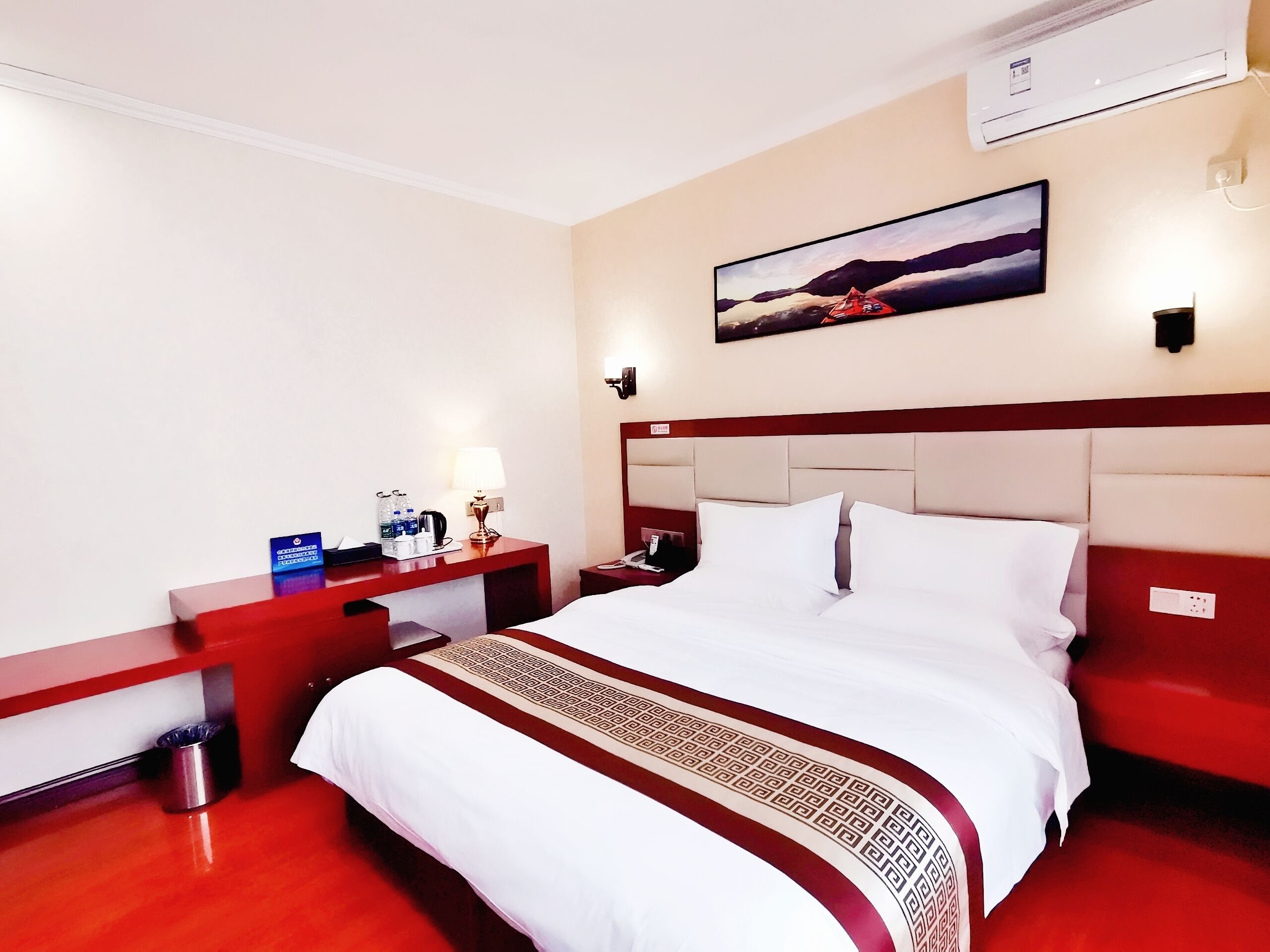 Quarto de hotel com uma cama grande, uma mesinha de cabeceira, ar condicionado fixado na parede e um quadro acima da cama.