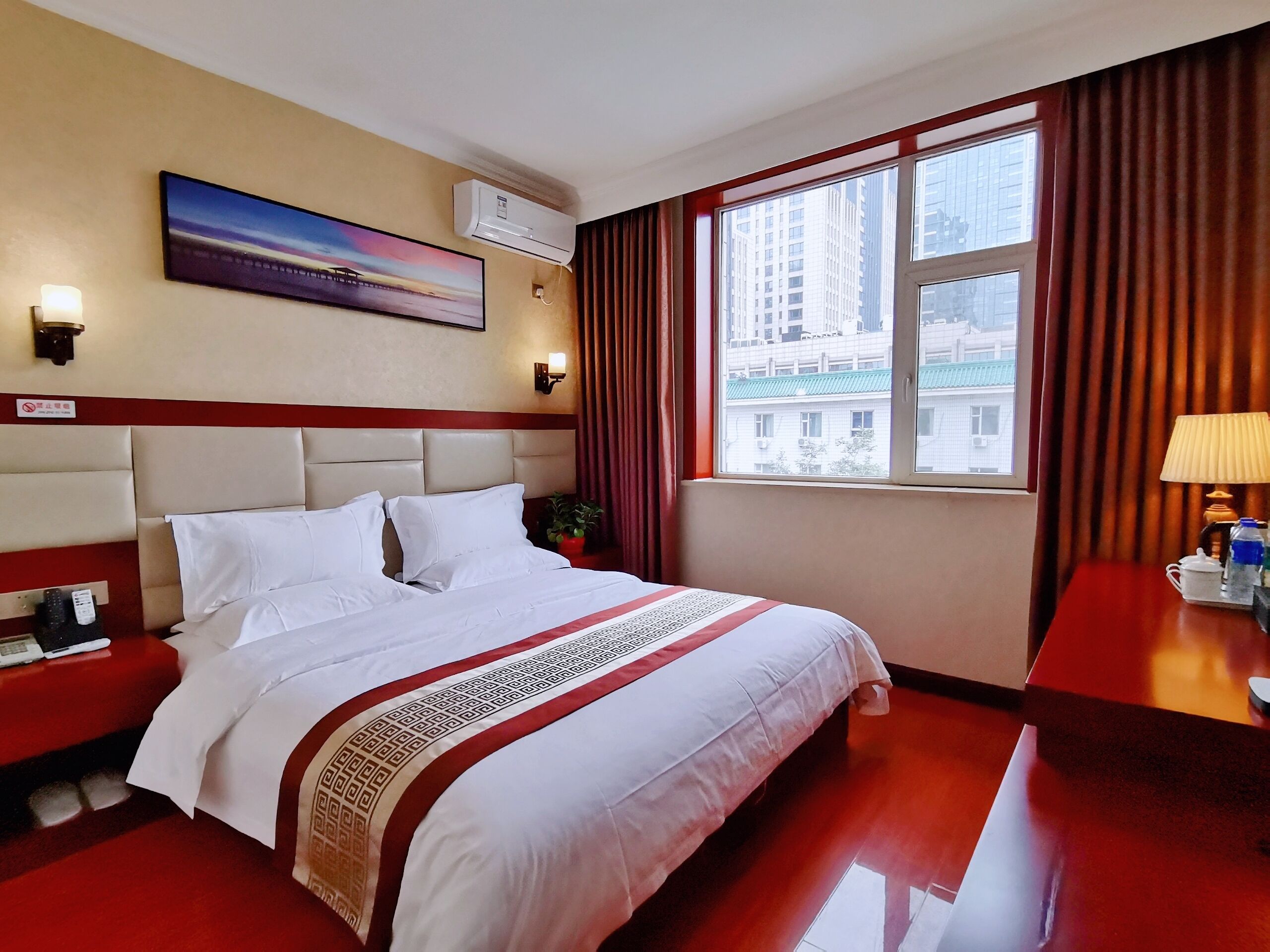 A hotel room with a large bed, a nightstand, a desk with a lamp, a window with curtains, a painting on the wall, and an air conditioning unit.