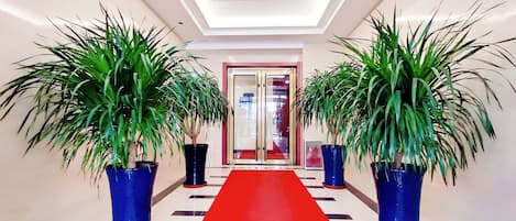A hotel corridor with a red carpet, potted plants, and a glass door entrance.