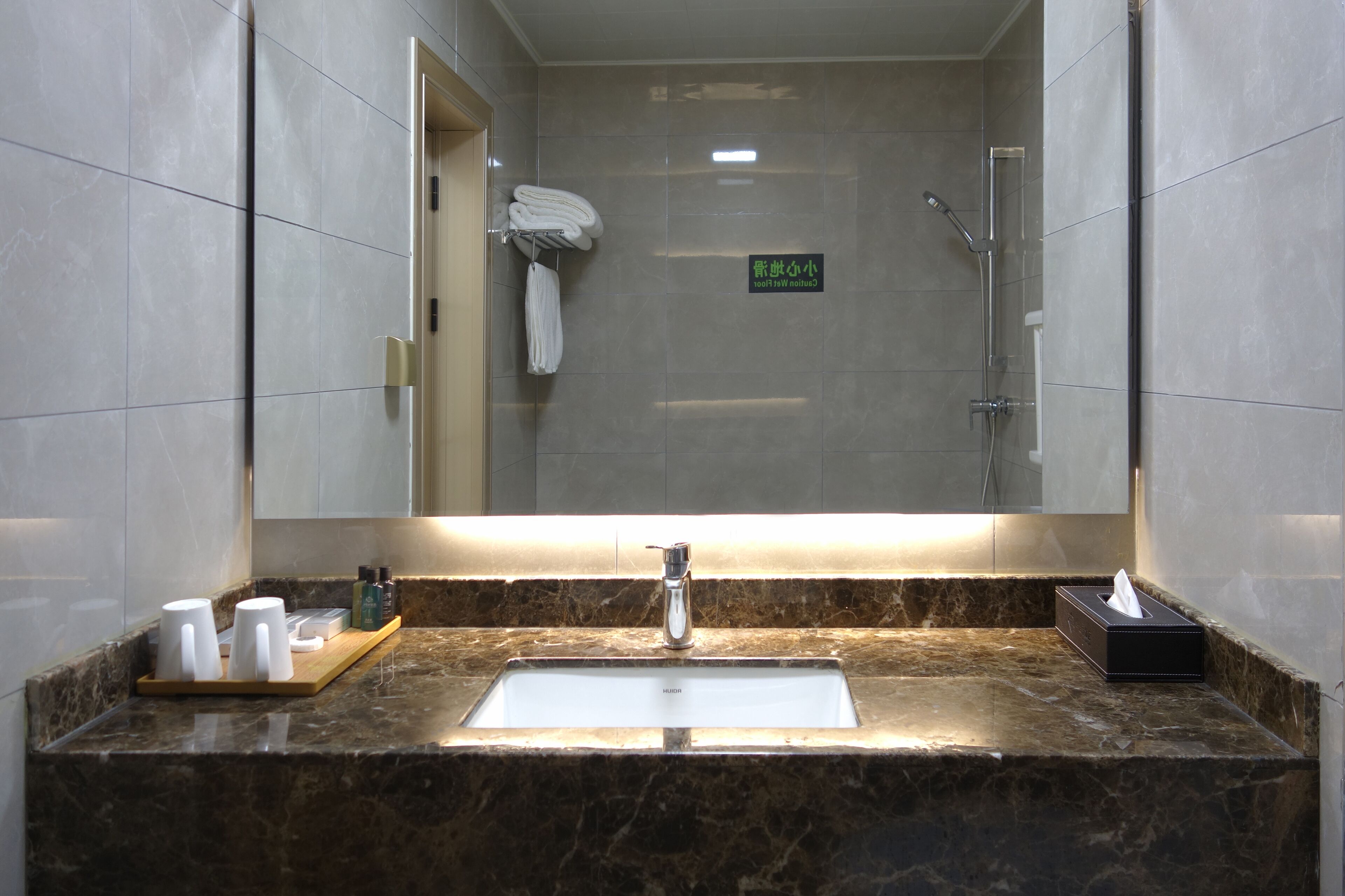 A bathroom with a large mirror, a sink, and a shower. There are towels hanging on a rack and a sign in the mirror.