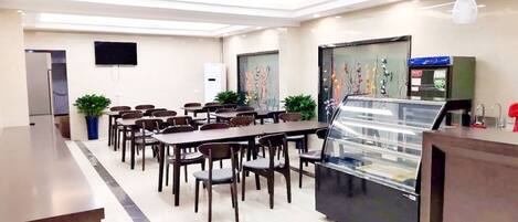 Un espace repas moderne avec plusieurs tables et chaises, un comptoir central doté d’une vitrine, et un téléviseur à écran plat fixé au mur.