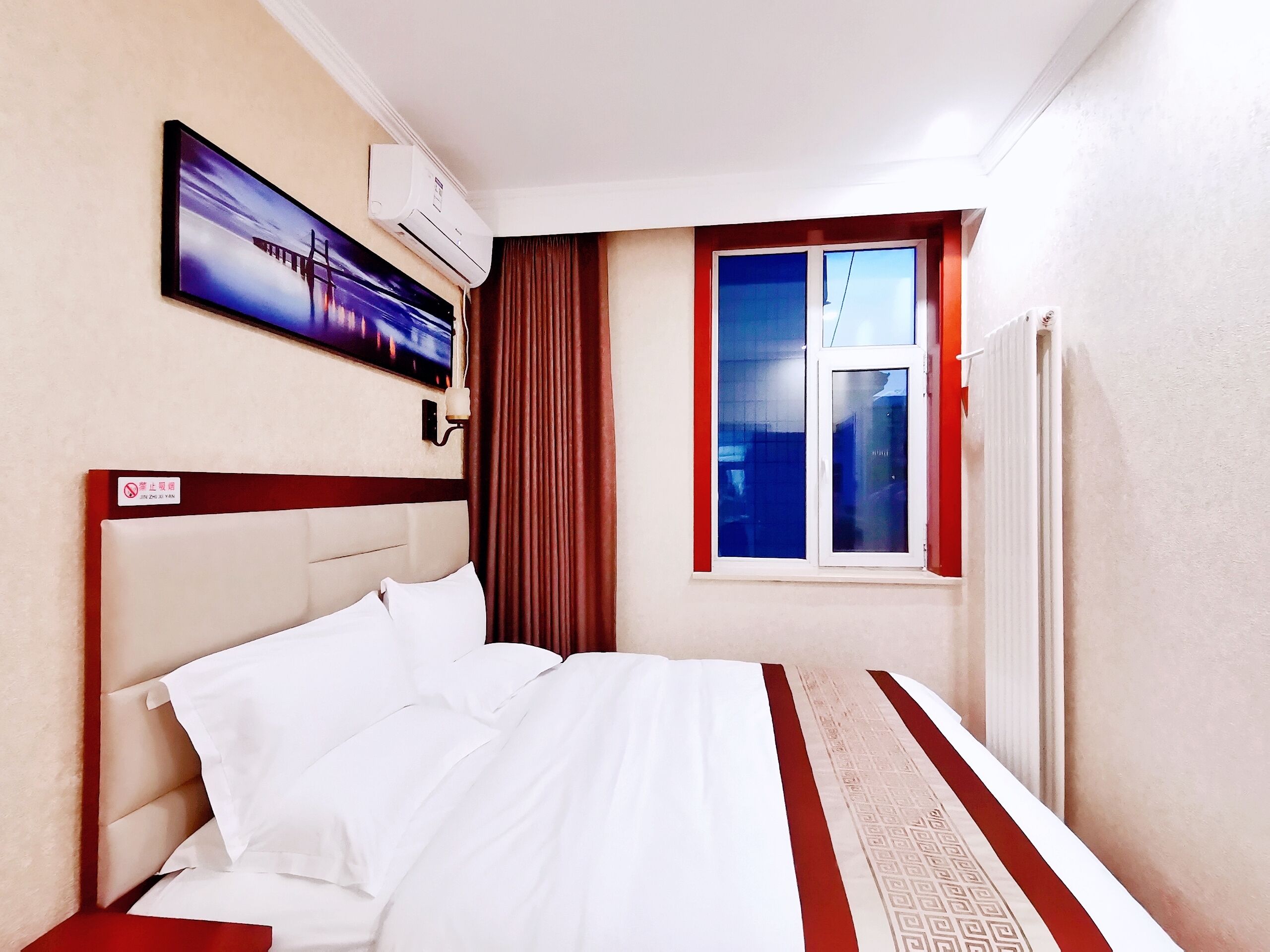 Ein Hotelzimmer mit Bett, Wandklimaanlage, Vorhängen am Fenster und einem Gemälde über dem Bett.