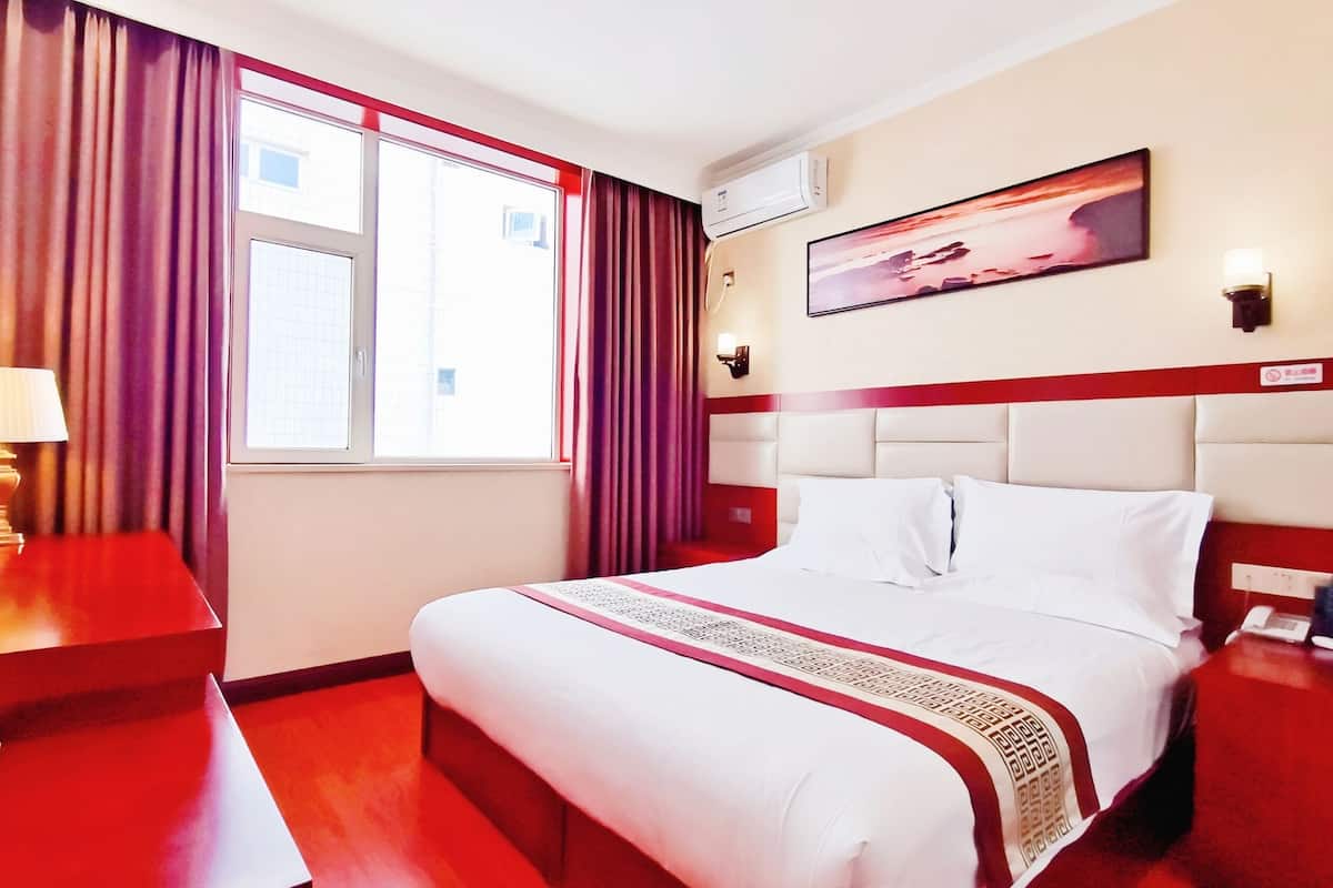Une chambre d’hôtel avec un grand lit, des rideaux rouges, une fenêtre donnant sur un beau paysage, un appareil de climatisation, un tableau au mur et une table de chevet avec une lampe.