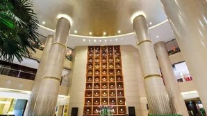 Um hall amplo com colunas imponentes, uma parede decorativa central com padrões circulares e uma fonte com flores.