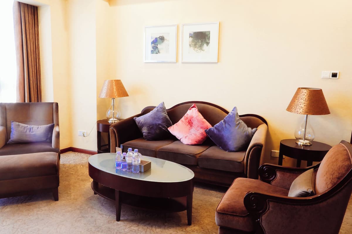 Ein Wohnzimmer mit einer braunen Couch, zwei Sesseln, einem Couchtisch und zwei Lampen. An der Wand hängen zwei gerahmte Bilder.