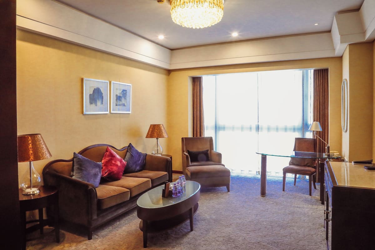 Ein Hotelzimmer mit einer Couch, einem Sessel und einem Glastisch. Es gibt einen Schreibtisch mit Lampe und Stuhl. Das Zimmer verfügt über ein großes Fenster mit Vorhängen.