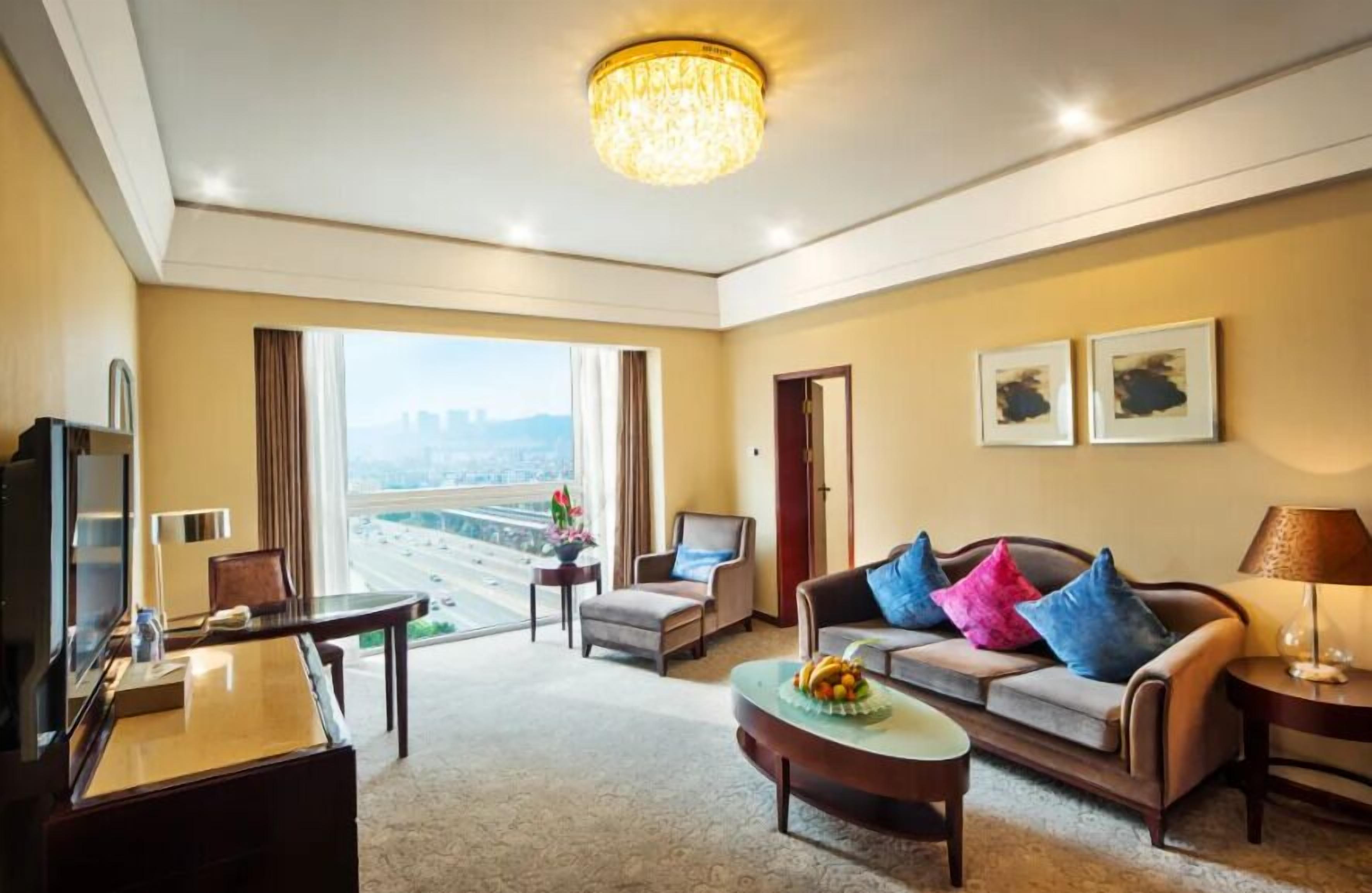 A hotel room with a large window, a sofa with colorful cushions, a desk with a computer, and a chandelier.