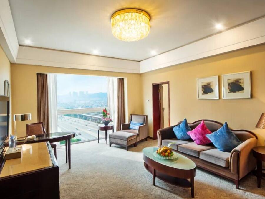 A hotel room with a large window, a sofa with colorful cushions, a desk with a computer, and a chandelier.