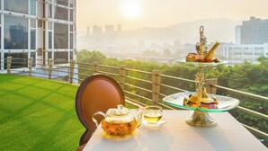 Uma mesa com um bule e xícara, um suporte para sobremesas de três andares e uma vista de uma paisagem urbana com vegetação ao fundo.