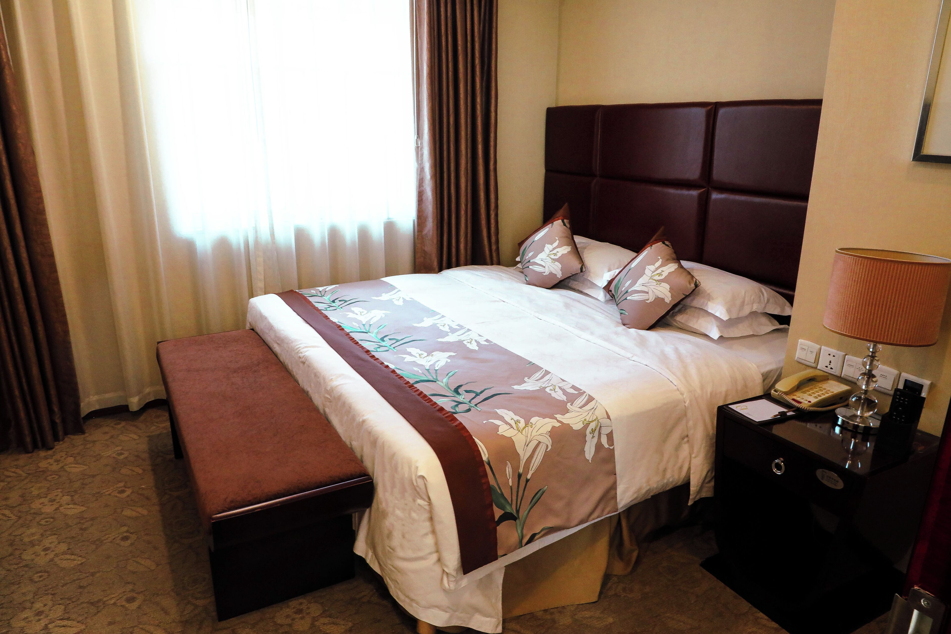 Cama bem arrumada com colcha florida, um banco marrom e uma mesinha de cabeceira com luminária e telefone.