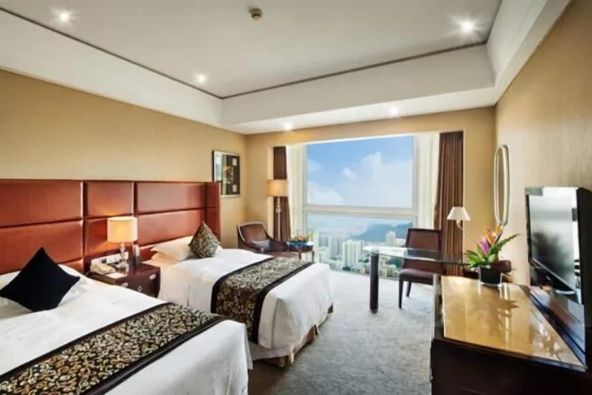Ein Hotelzimmer mit zwei Betten, einem Schreibtisch, einem Fernseher und einem großen Fenster mit Blick auf die Stadt.