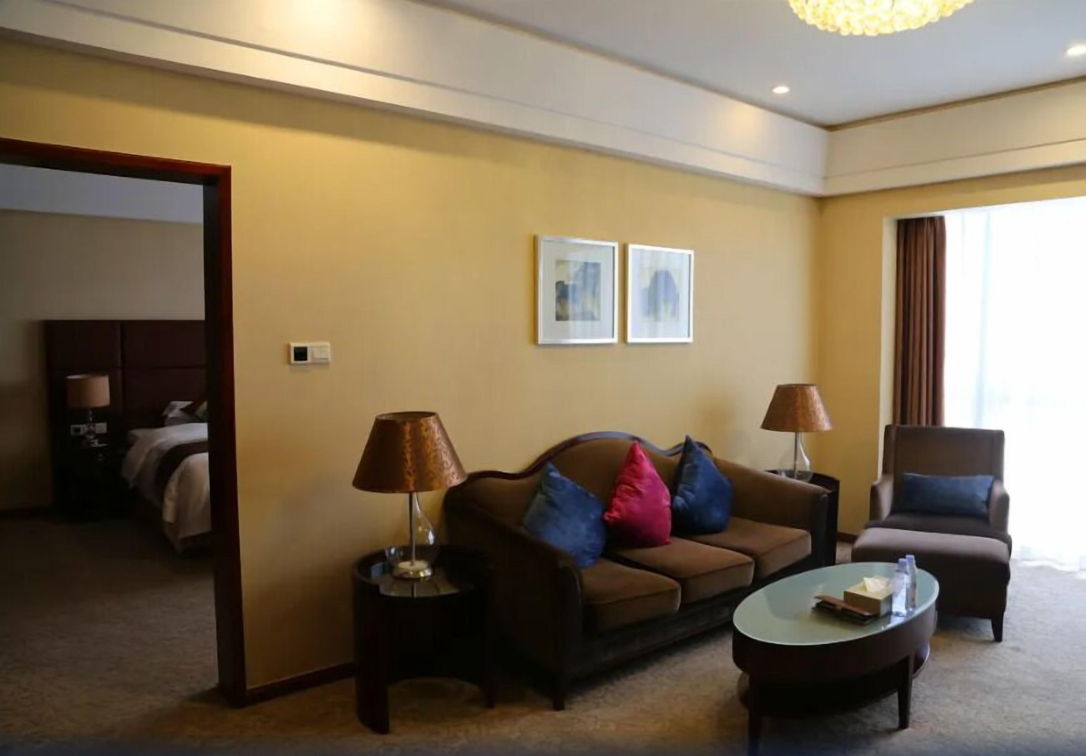A hotel room with a sofa, armchair, coffee table, and two lamps. There is a bed in the background and two framed pictures on the wall.