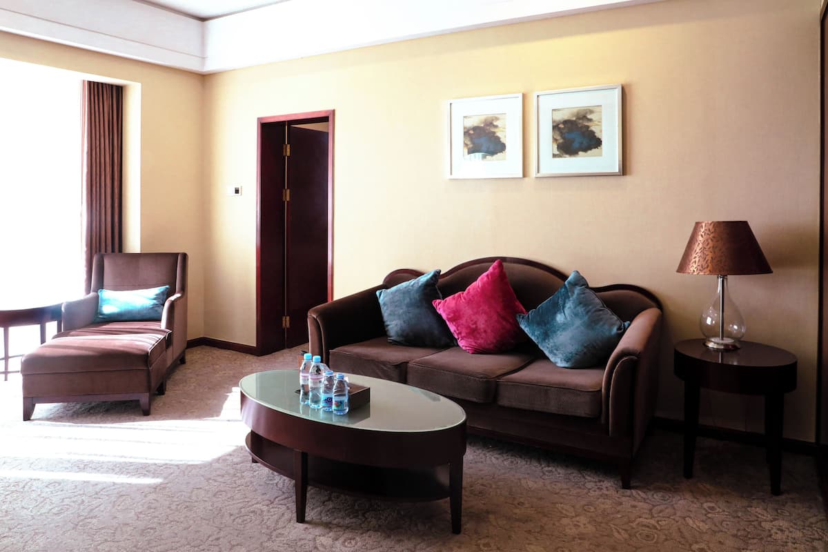 Ein Hotelzimmer mit braunem Ledersofa, einem braunen Sessel, einem Glastisch mit Wasserflaschen, einer Lampe auf einem Beistelltisch und zwei gerahmten Kunstwerken an der Wand.