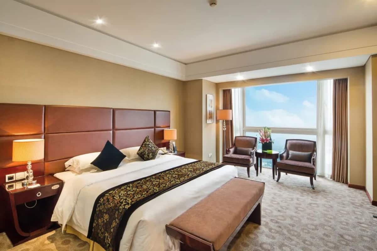 Ein Hotelzimmer mit einem großen Bett, einer Bank, zwei Sesseln, einem kleinen Tisch mit einer Lampe und einem großen Fenster mit Blick.