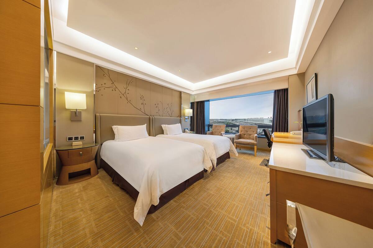 Ein Hotelzimmer mit zwei Betten, einem Schreibtisch, einem Fernseher und Blick auf die Stadt.