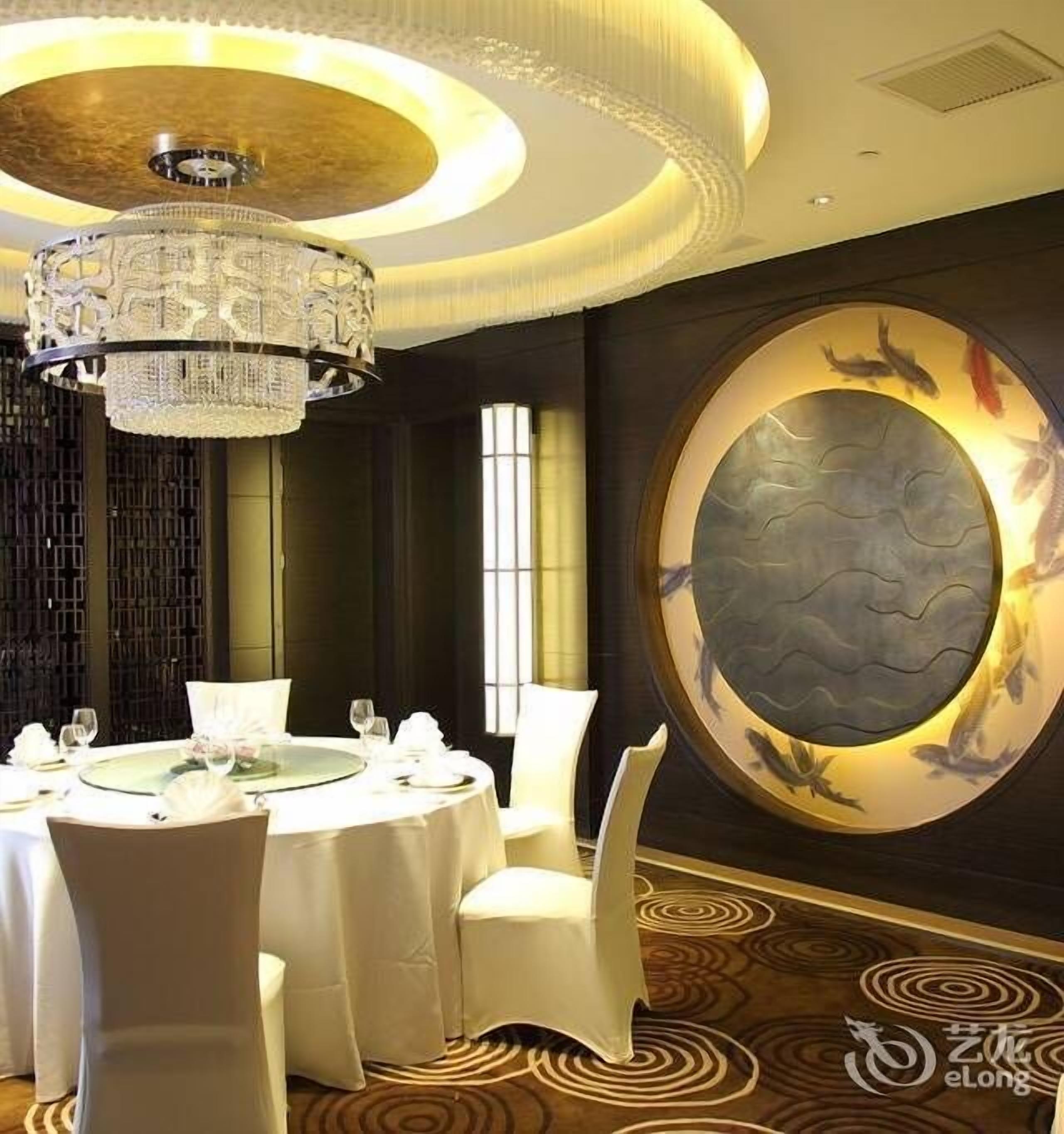 A dining area with a round table set for a meal, surrounded by white chairs, a large chandelier, and a decorative wall with a circular design.