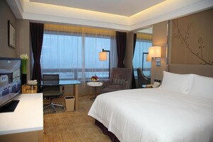 A hotel room with a large bed, a desk with a computer, a chair, a small table with a lamp, and a large window with curtains.