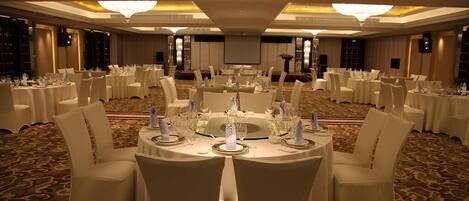 A banquet hall with round tables set for dining, white chairs, and a carpeted floor.
