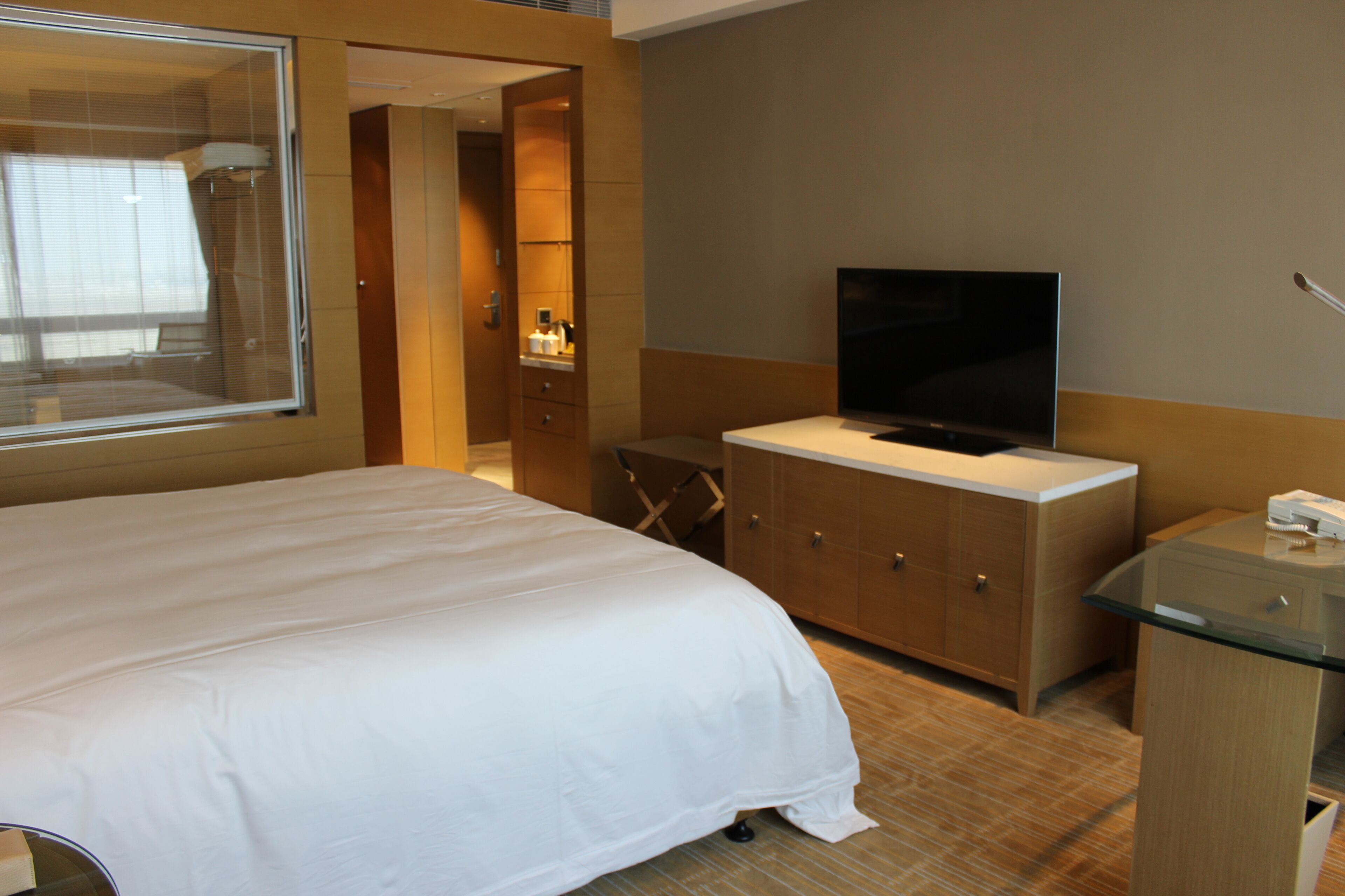 A hotel room with a large bed, a TV, a glass cabinet, and a bathroom visible through an open door.