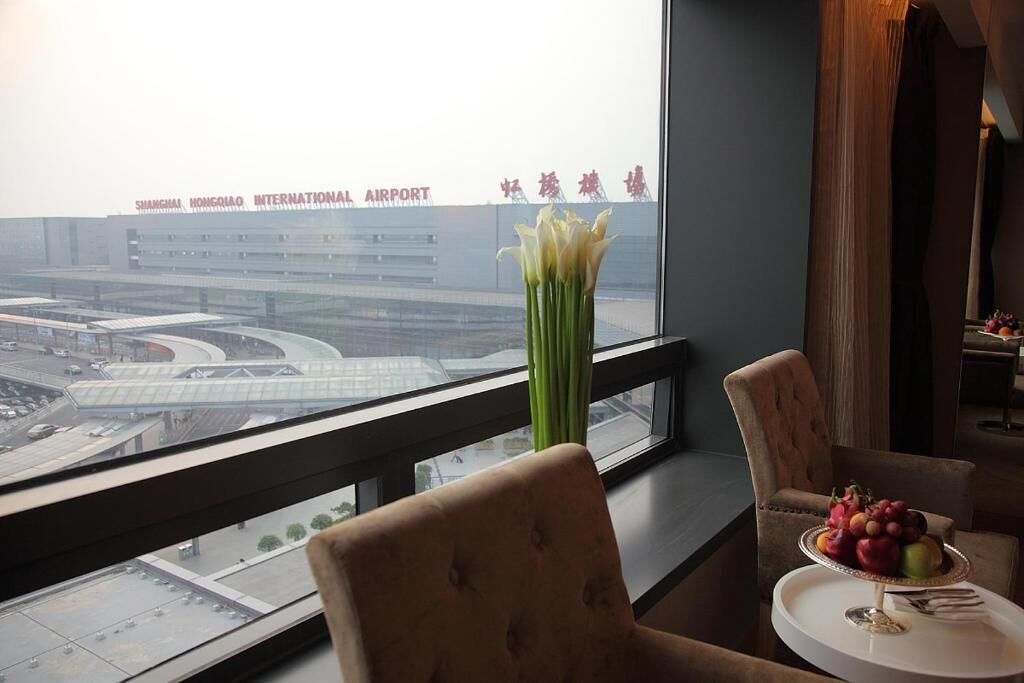 Een hotelkamer met uitzicht op een vliegveld, een bloevas en een schaal fruit op de tafel.