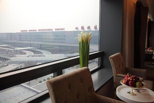 A hotel room with a view of an airport, a vase of flowers, and a fruit bowl on the table.