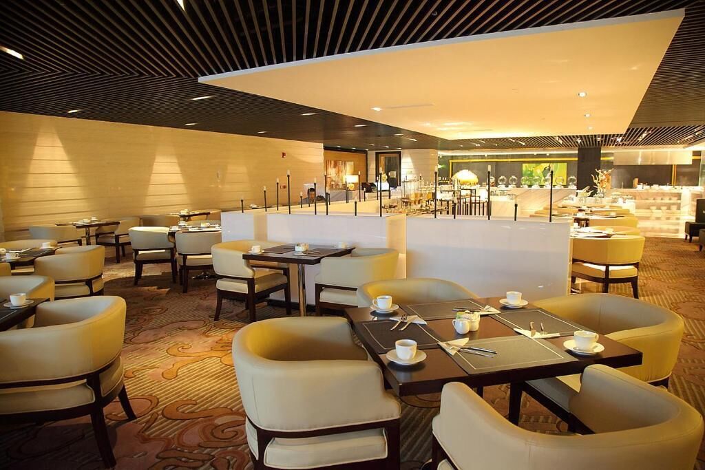 A modern hotel lobby with a central seating area, a reception desk, and a view of the restaurant in the background.