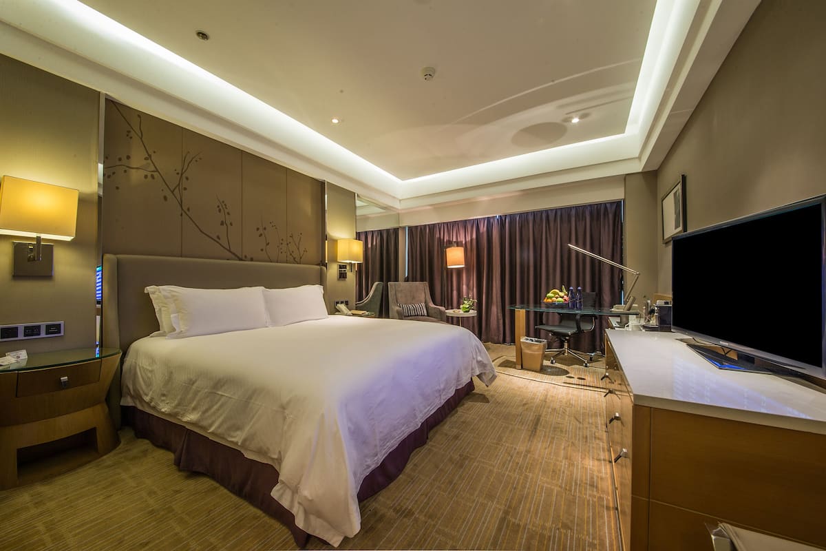 Ein Hotelzimmer mit einem großen Bett, einem Schreibtisch, einem Flachbildfernseher, einem Sessel, einer Lampe und einem Spiegel.