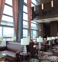A modern hotel lobby with leather sofas, round tables with lamps, large windows, and a high ceiling with vertical wooden slats.