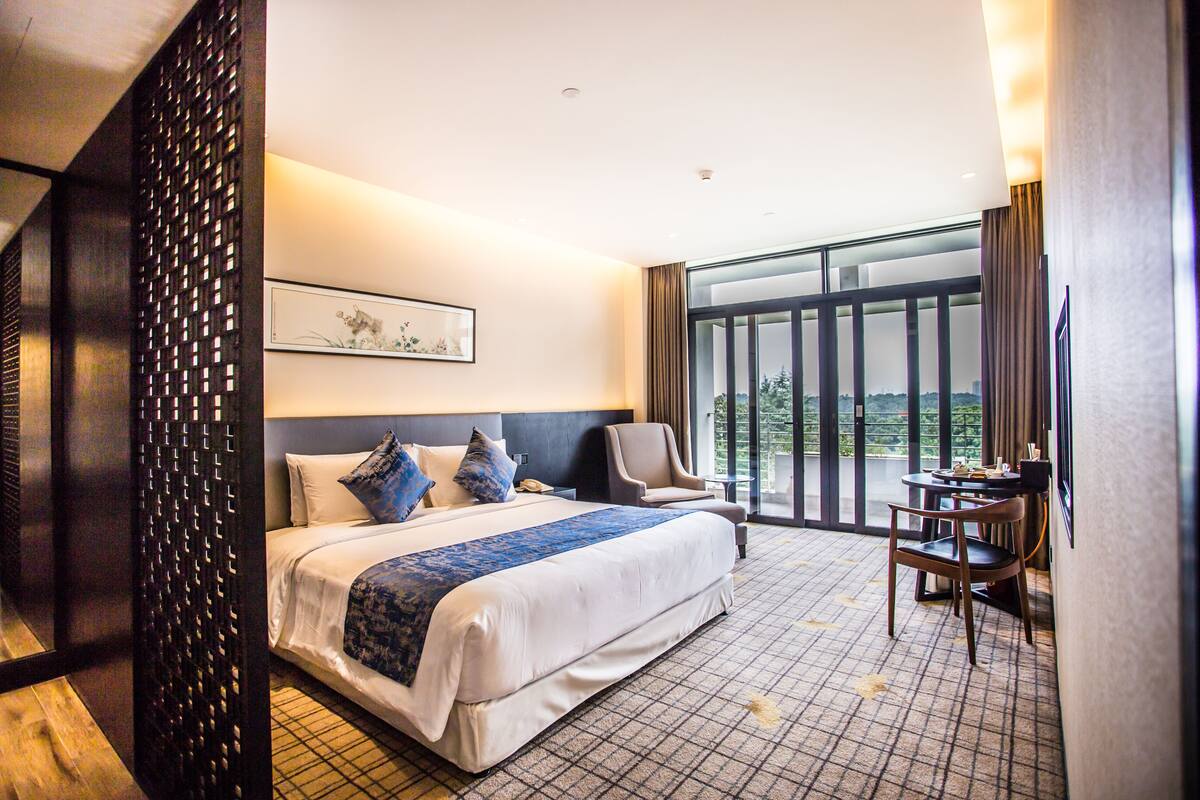 A hotel room with a large bed, a chair, a small table, and a view of the outdoors through a large window.