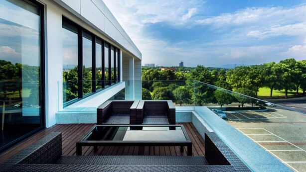 A modern outdoor balcony with a glass railing, wicker furniture, and a view of a parking lot and greenery. - Preess Resort Hotel (Changsha)