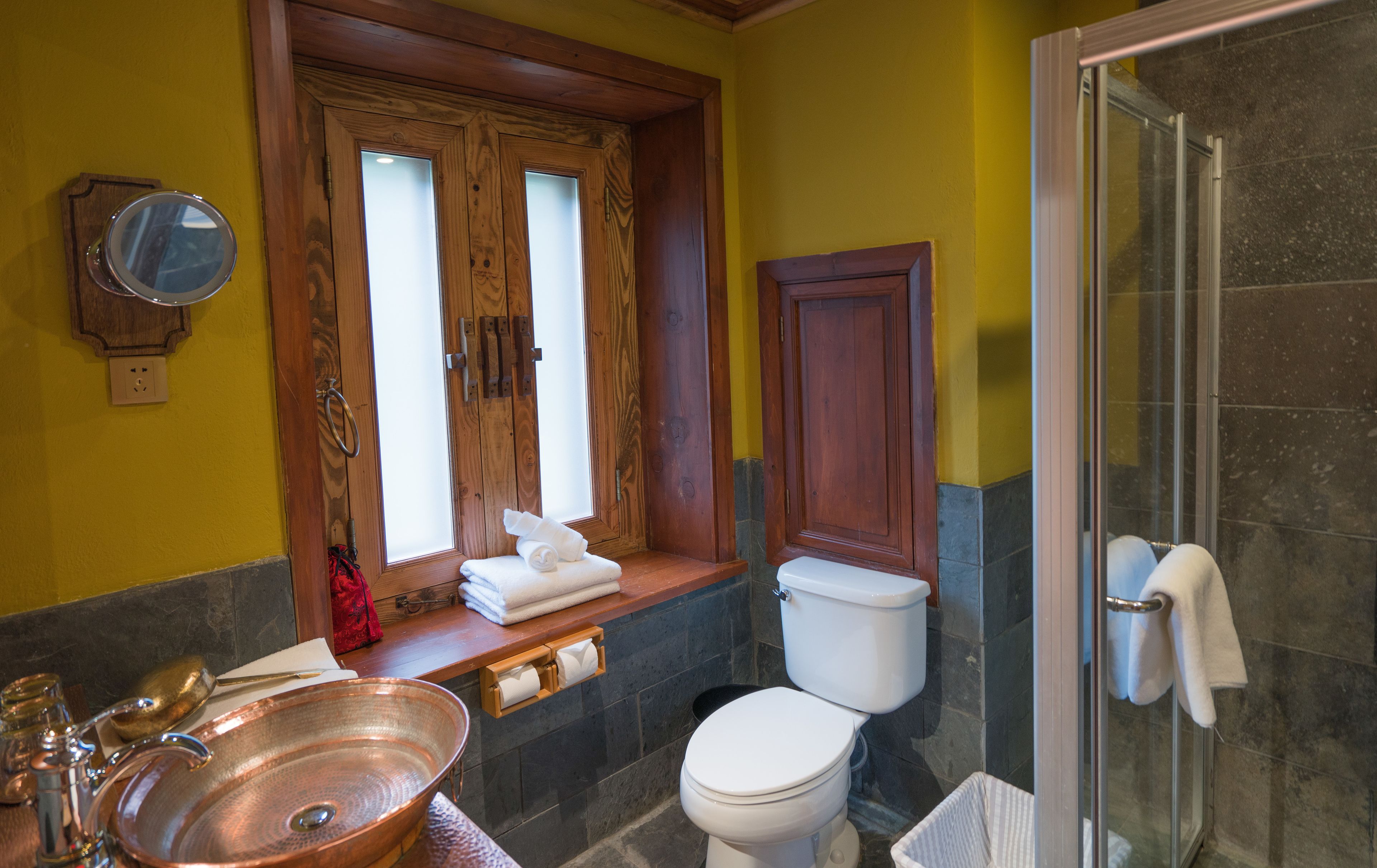 A bathroom with a wooden vanity, a copper basin, a toilet, and a glass shower enclosure.