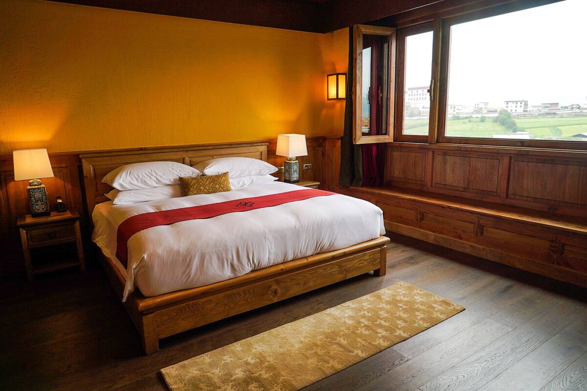 A large bed with a wooden frame, two bedside tables with lamps, a red throw blanket, and a view of buildings through a window.