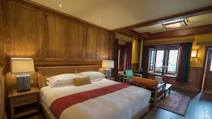 A large bed with a red runner, wooden paneling, a nightstand with a lamp, and a view of the city through the window. - Songtsam Linka Shangri-La (Deqin)
