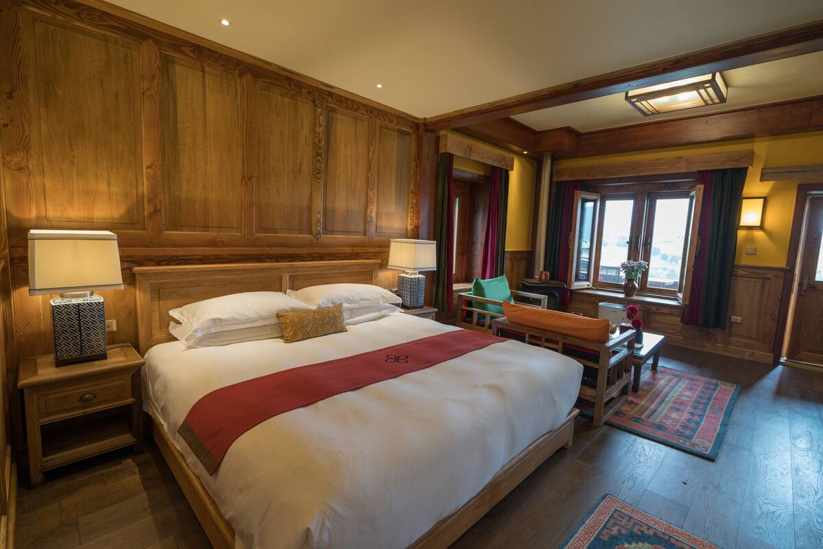 A large bed with a red runner, wooden paneling, a nightstand with a lamp, and a view of the city through the window.