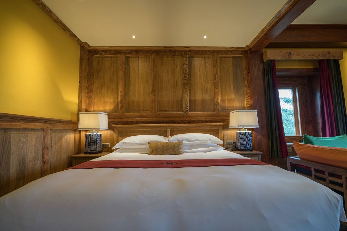 A large bed with white linens, two bedside tables with lamps, wooden paneling on the walls, and a window with curtains.