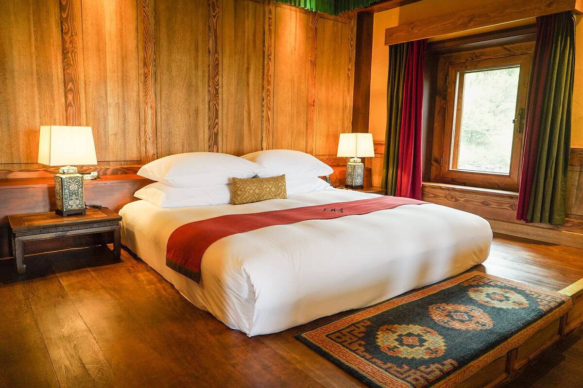 A large bed with white linens, two bedside tables with lamps, a wooden headboard, and a window with curtains.